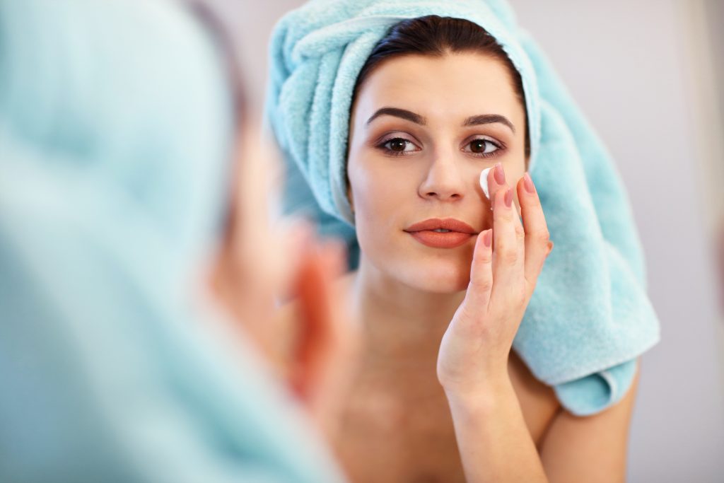 Young woman standing in bathroom and applying face cream in the morning
