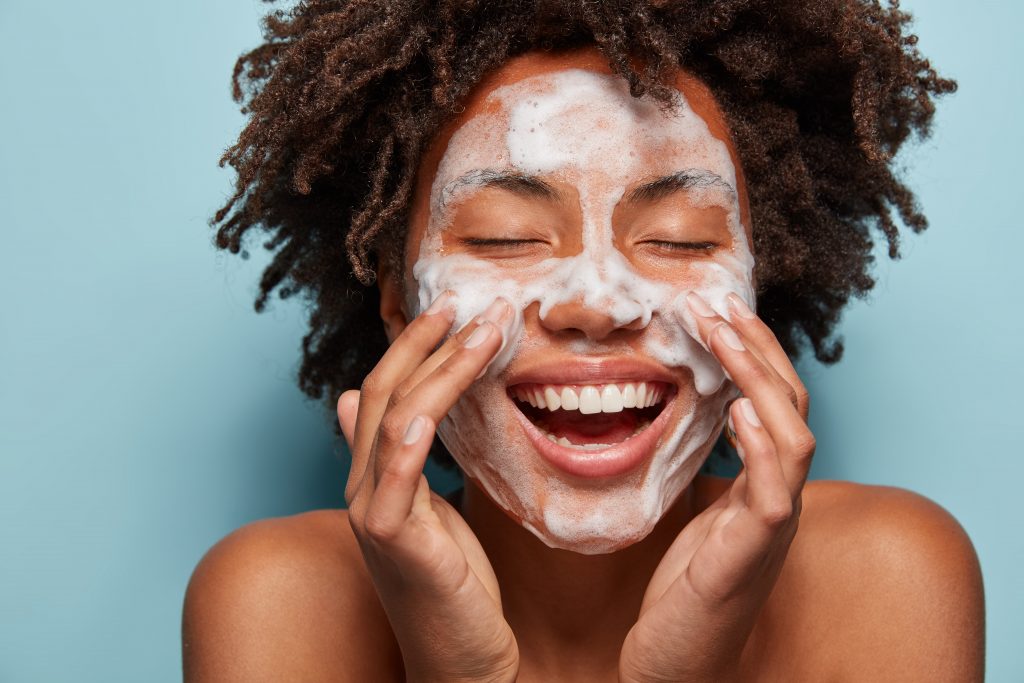 Spa, therapy and beauty concept. Satisfied black woman washes face with white soap, has cleansing foam, purification of face, cleans pores, shows bare shoulders, isolated over blue studio wall