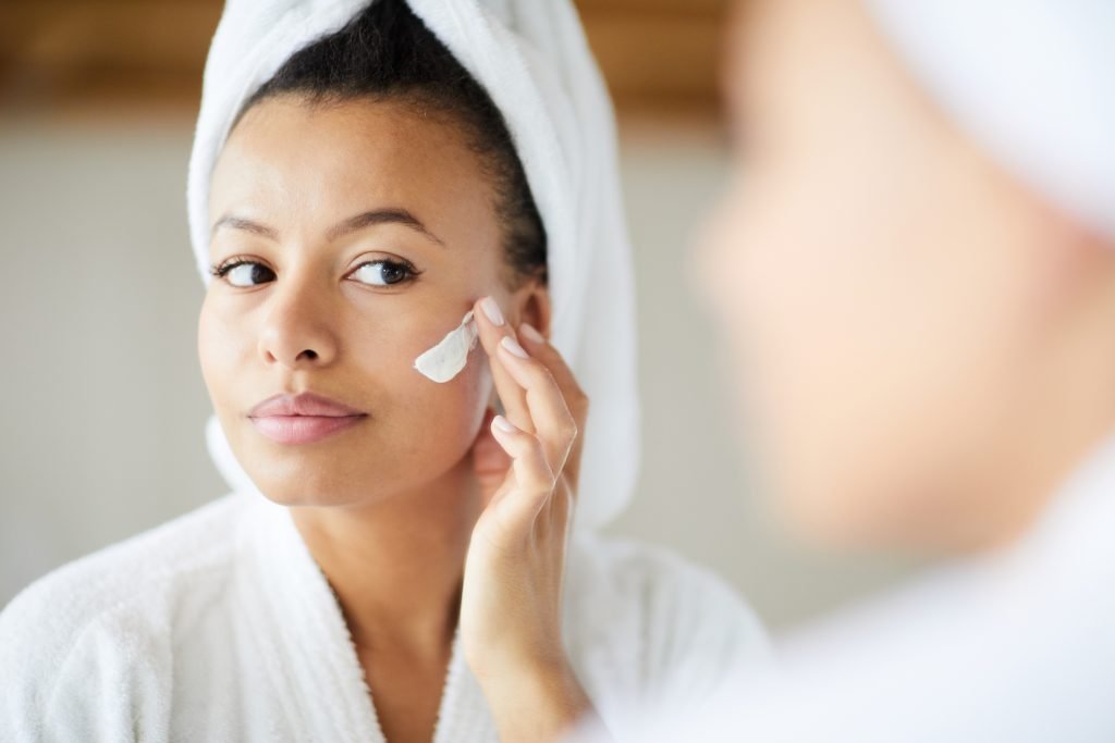 Head and shoulders portrait of beautiful Mixed-Race woman applying face cream during morning routine, copy space
