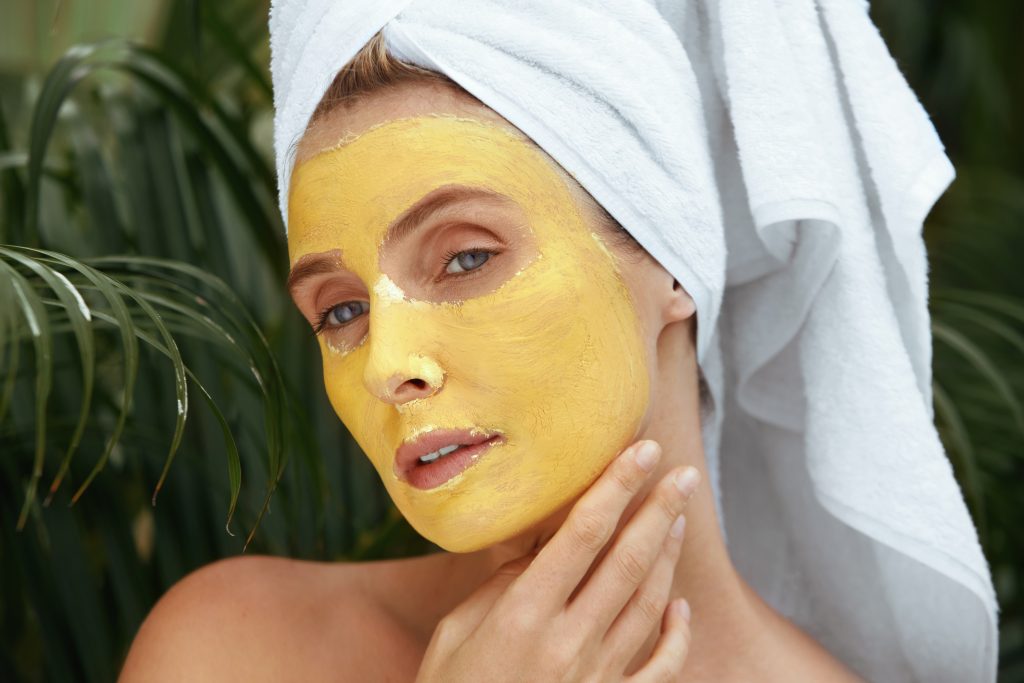 Beauty Mask. Woman In Bath Towel On SPA Procedure Close Up Portrait. Face Covered With Yellow Skin Care Product For Oil Derma. Anti-Aging Therapy At Tropical Resort.