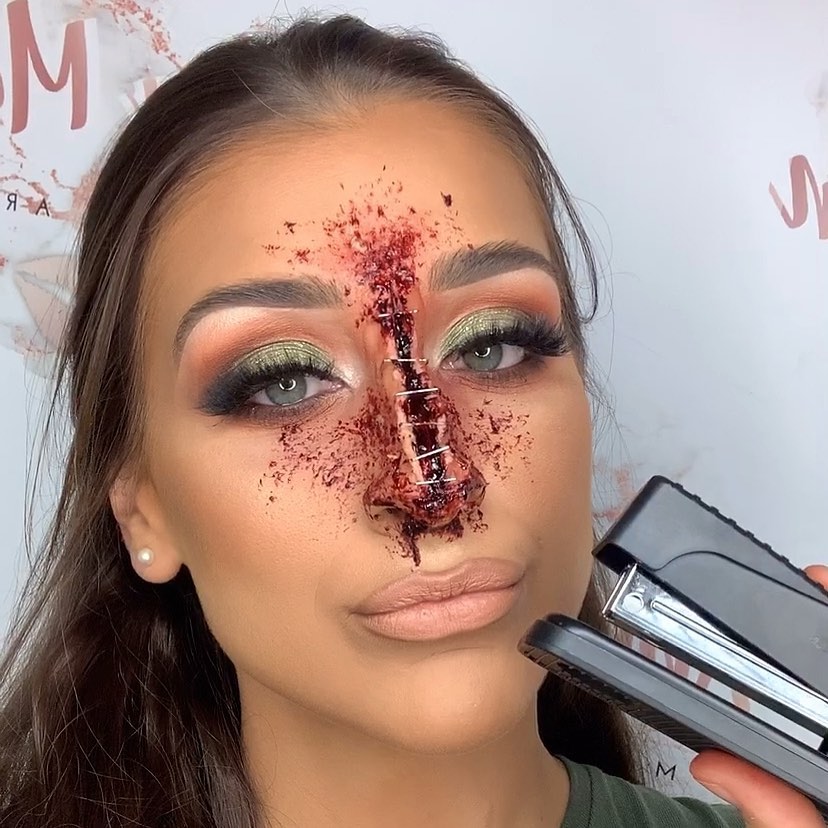 A person with a bloody makeup effect showing a stapled nose, holding a stapler. The rest of the makeup is glamorous, with green eyeshadow and nude lips, blending beauty with horror.