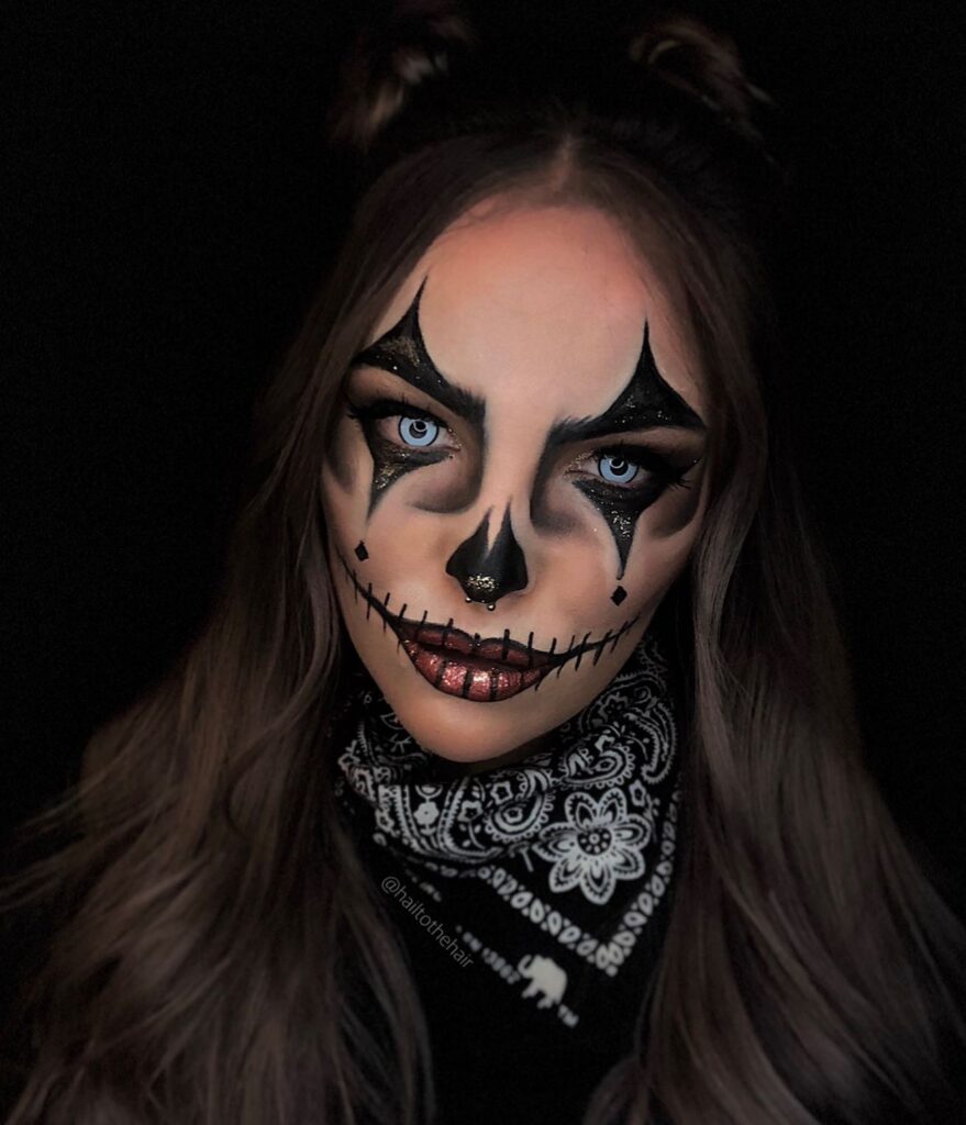 Person wearing gothic skull-inspired Halloween makeup with bold black and gold accents, metallic lips, white contact lenses, and a stitched mouth design, styled for a haunting yet glamorous effect.