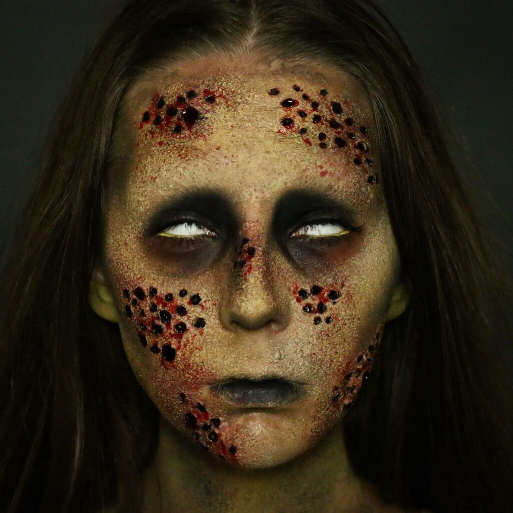 A person with zombie makeup featuring sunken blackened eyes, a pale complexion, and bloodied, festering wounds across the face, giving a terrifying infected appearance.