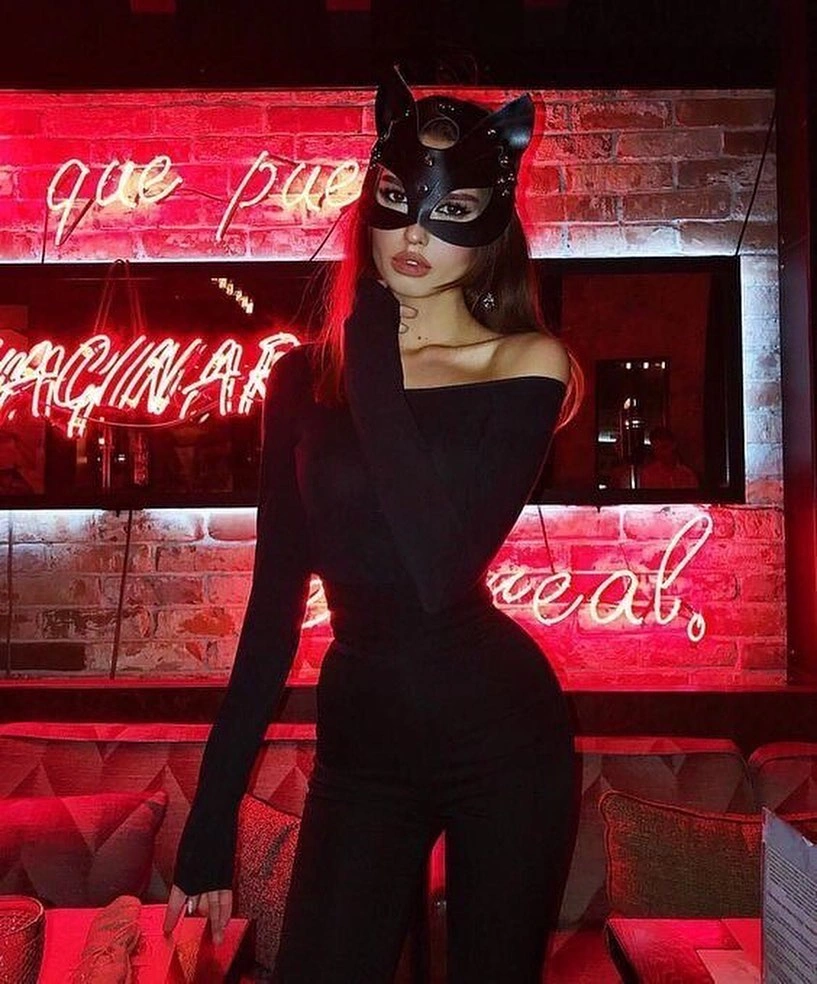 Woman dressed in an all-black outfit with a cat mask, standing in front of a neon-lit wall.