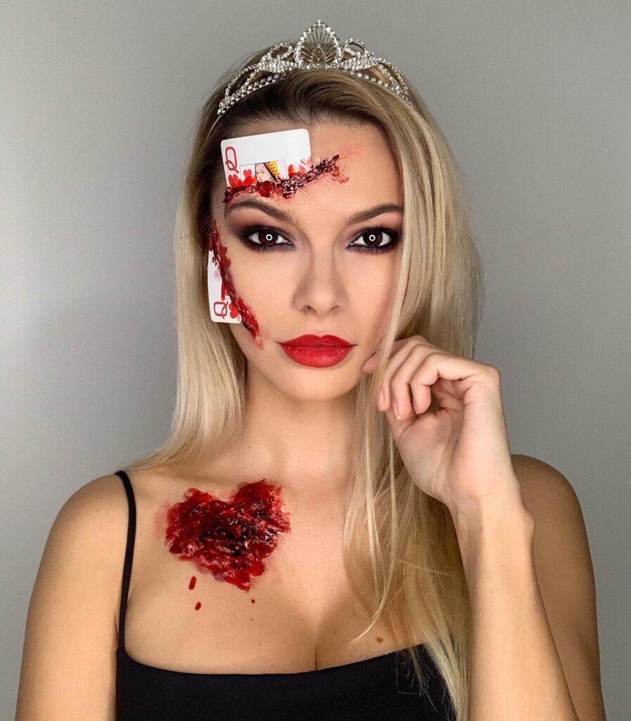 Person wearing Queen of Hearts-themed Halloween makeup with playing cards embedded in bloody wounds, a tiara, red lipstick, and dramatic eye makeup.