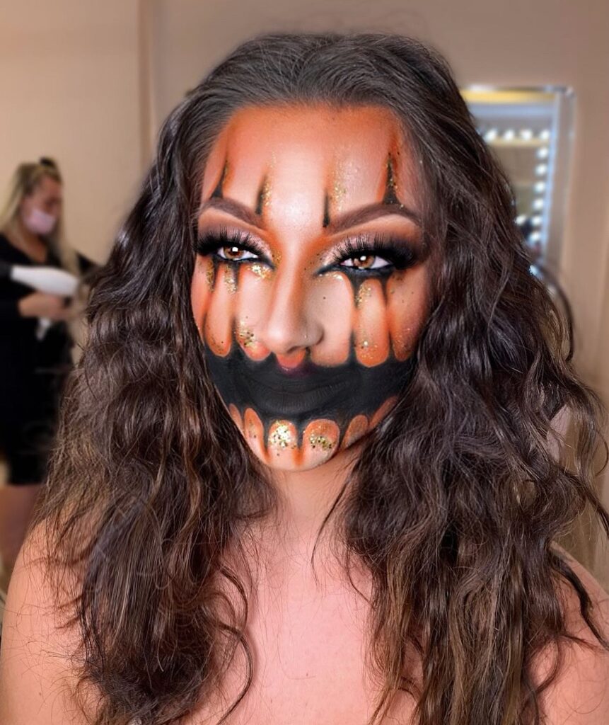 A person with pumpkin-themed makeup, featuring orange and black shading, glitter accents, and a sharp, glowing grin for a glamorous Halloween look.