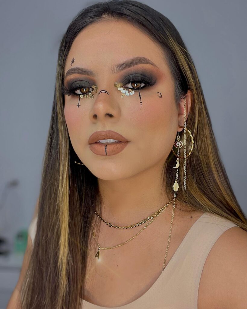 Woman with dark smoky eye makeup featuring golden glitter tears, subtle face line details, and nude lips. She accessorizes with layered gold jewelry and celestial-themed earrings.