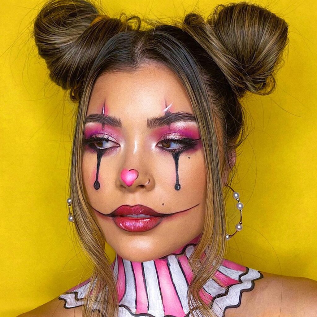 A woman with pink and black clown makeup, featuring black teardrops under the eyes, a pink heart-shaped nose, and a striped collar.