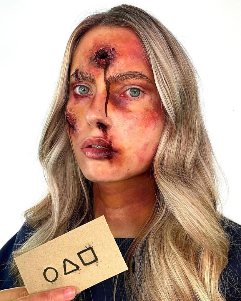 Person wearing Squid Game-inspired Halloween makeup with realistic bullet wounds, bruising, blood effects, and holding a card with Squid Game symbols for a brutalized look.