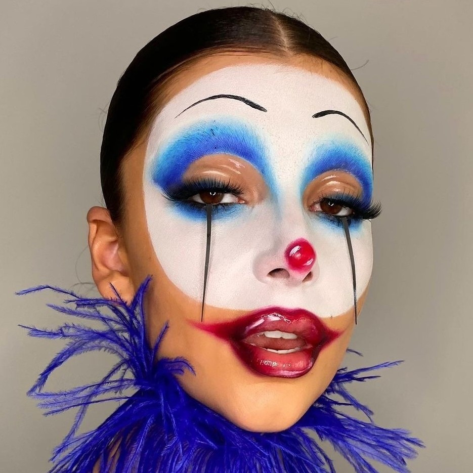 A person with Pierrot-style clown makeup, characterized by blue eyeshadow, a red nose, glossy lips, and black tears on a white base, wearing a vibrant blue feather collar.