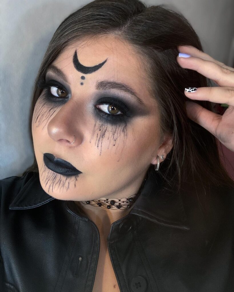 A person with gothic makeup, featuring a crescent moon on the forehead, smudged black eyeshadow, and bold black lipstick, wearing a leather jacket and choker.