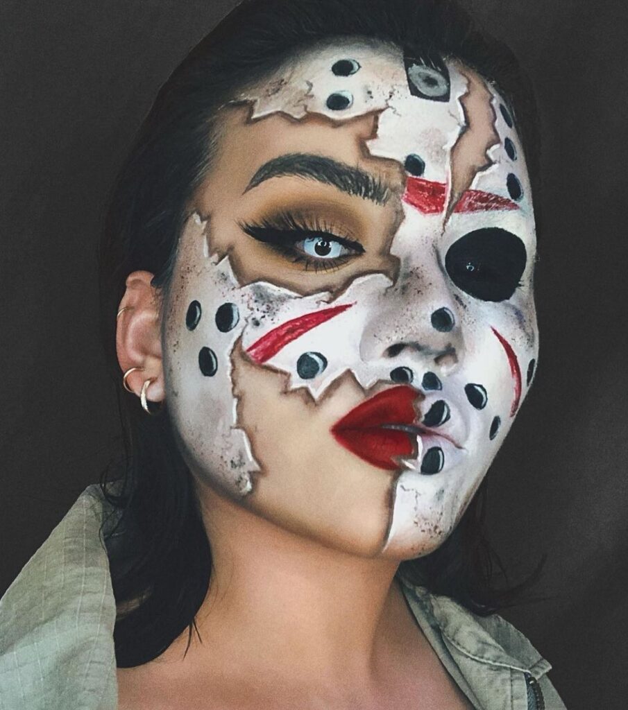 A woman wearing makeup resembling a cracked and torn Jason Voorhees mask with red accents and dark eye patches, creating a haunting horror look.