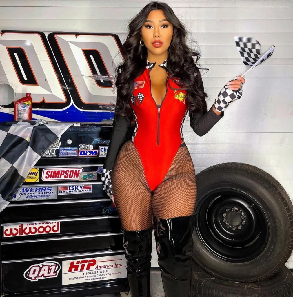 A woman dressed in a red racing bodysuit with checkered flag details, fishnet stockings, and thigh-high black boots, holding a small checkered flag, standing confidently next to racing equipment.