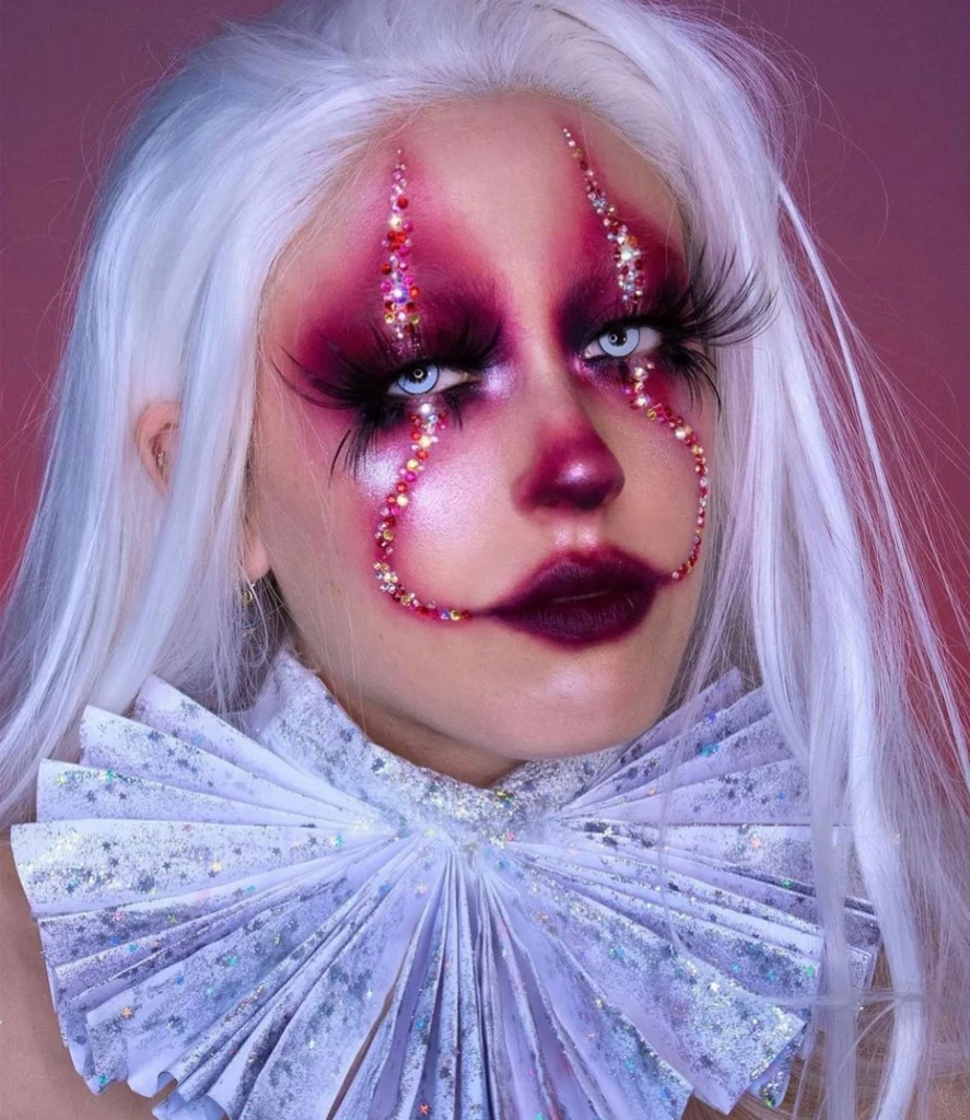 Close-up of a person with vibrant pink clown makeup, adorned with rhinestones and wearing a white wig and ruffled collar.