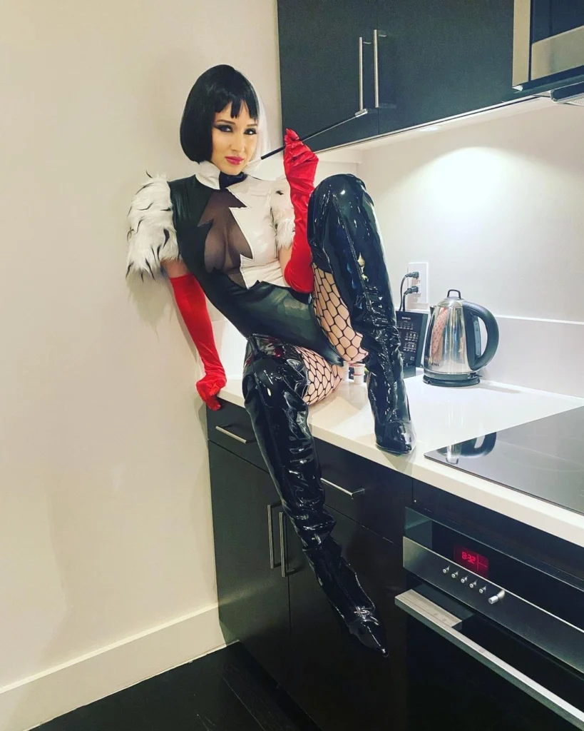 A person dressed as a stylish villainess, wearing a black bob wig, latex bodysuit with a white faux fur collar, red gloves, fishnet stockings, and thigh-high latex boots, holding a cigarette holder while seated on a kitchen countertop.