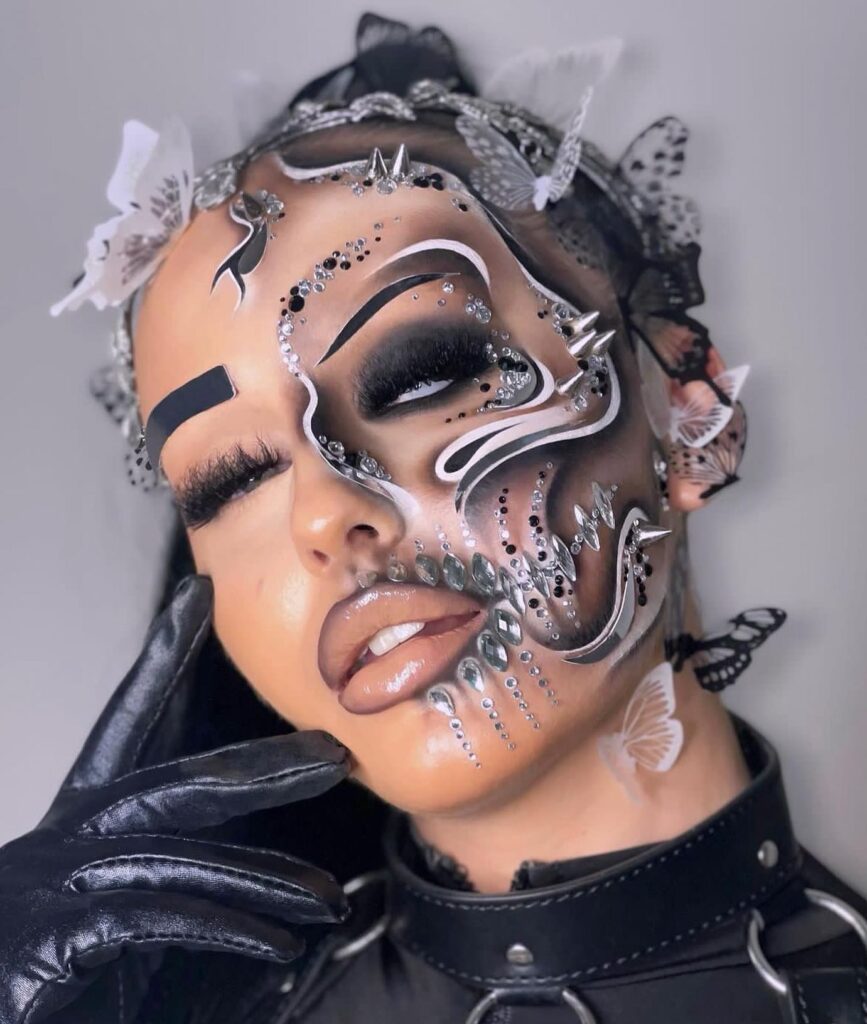 A person with futuristic makeup, featuring metallic details, rhinestones, bold black eyeshadow, and butterfly accents for a glamorous, ethereal look.