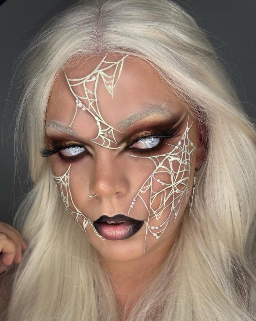 A person with platinum blonde hair wearing gothic-style makeup, including white spiderweb designs across the forehead and cheeks, smoky gold eyeshadow, black gradient lips, and white contact lenses.