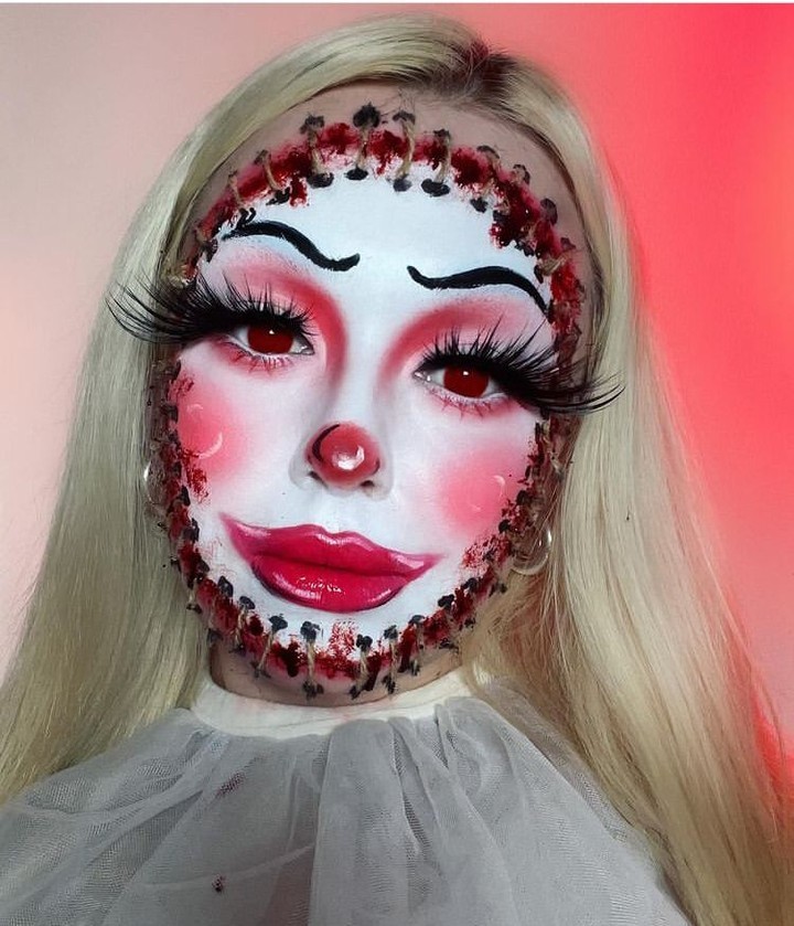 A person with doll-themed makeup, featuring red cheeks, lips, and nose, black stitches around the face, and exaggerated eyelashes for a creepy doll appearance.