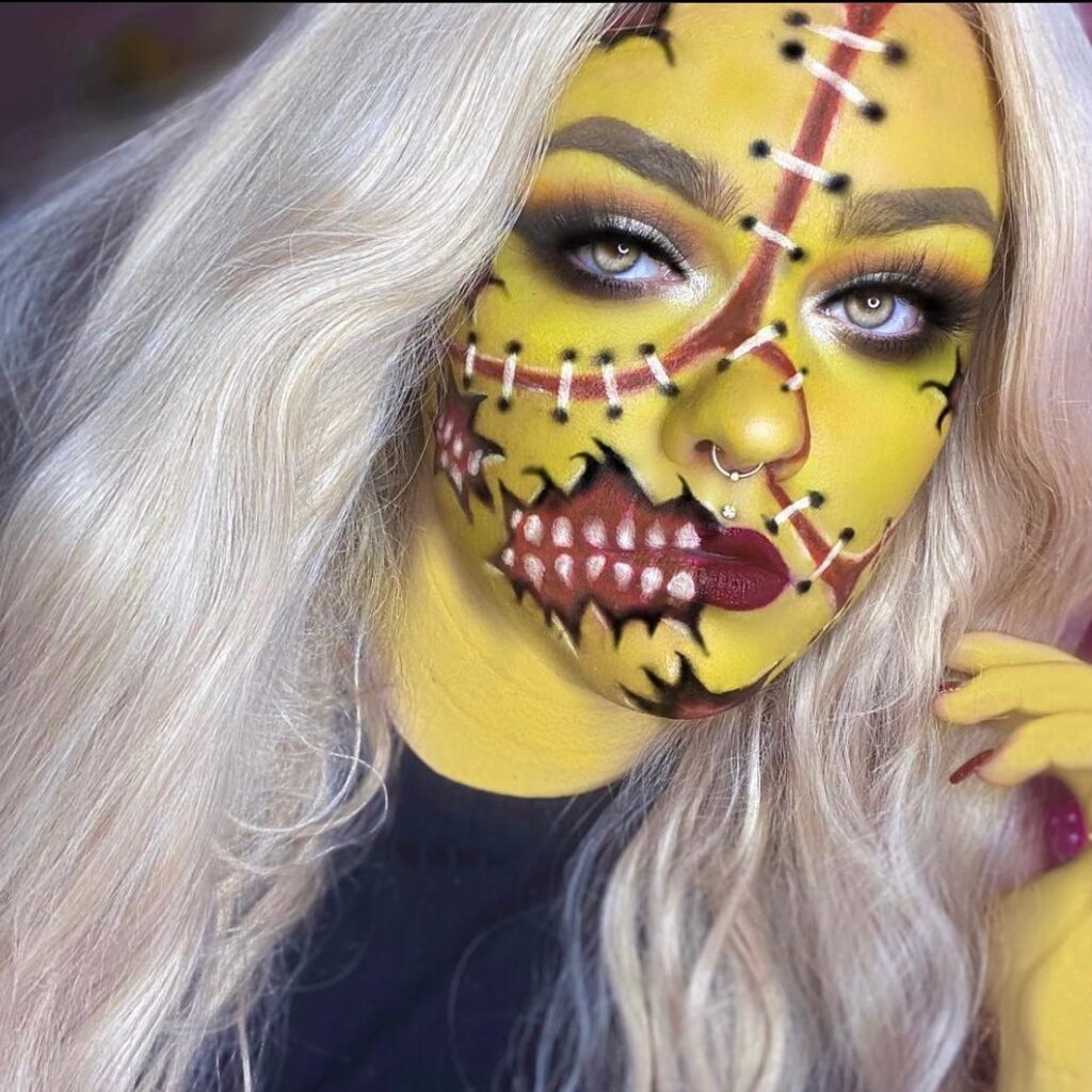 Person wearing stitched yellow Halloween makeup with exposed teeth, black stitching, smoky eyes, and silver hair for a creepy, horror-themed look.