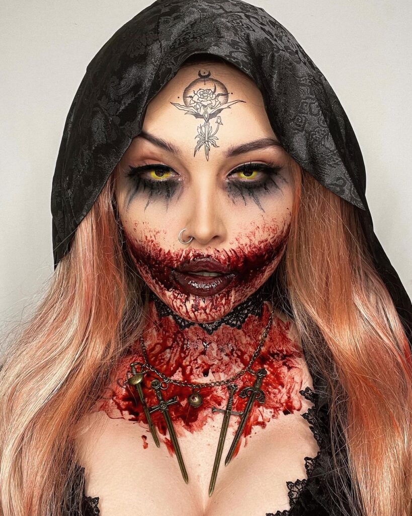 A person dressed as a vampire priestess with smeared blood around the mouth, yellow contacts, and a gothic tattoo on the forehead. The look is completed with a black hood and dagger necklace.