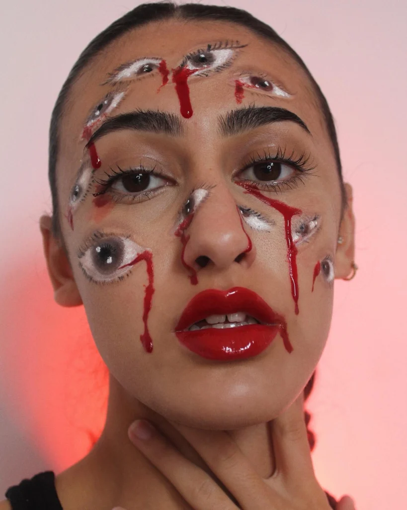 Close-up of a woman with Halloween makeup featuring multiple painted eyes across her face, each with blood-red tears, and glossy red lips. The overall effect is surreal and unsettling, creating a striking and eerie appearance.