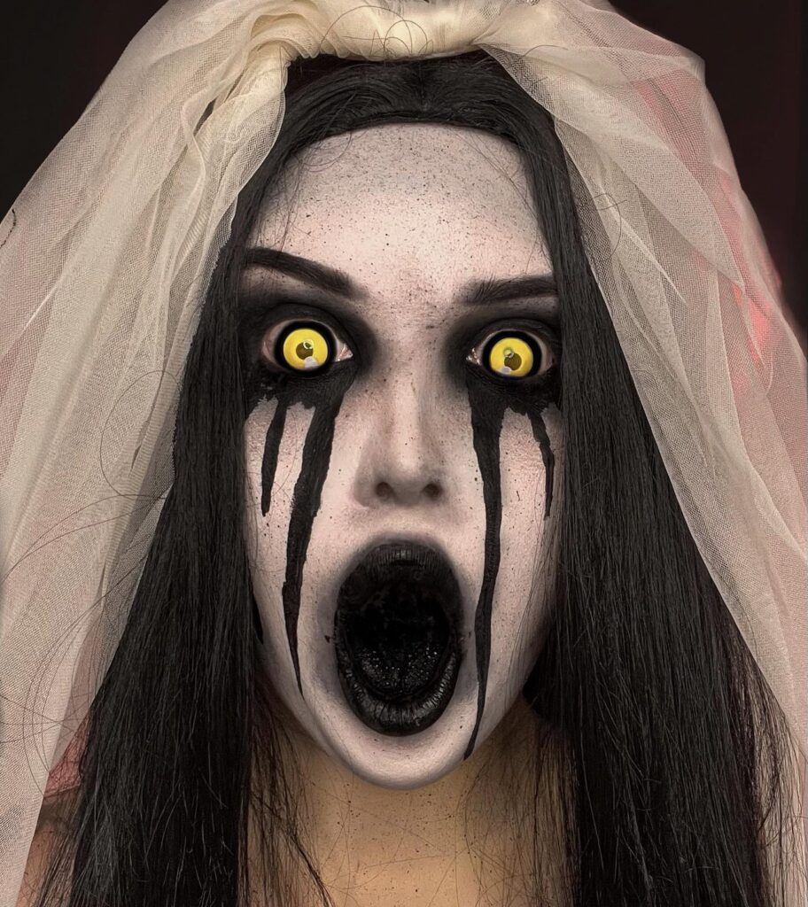 A person with ghost bride makeup, featuring white face paint, yellow eyes, black tears, and a gaping black mouth, wearing a veil.