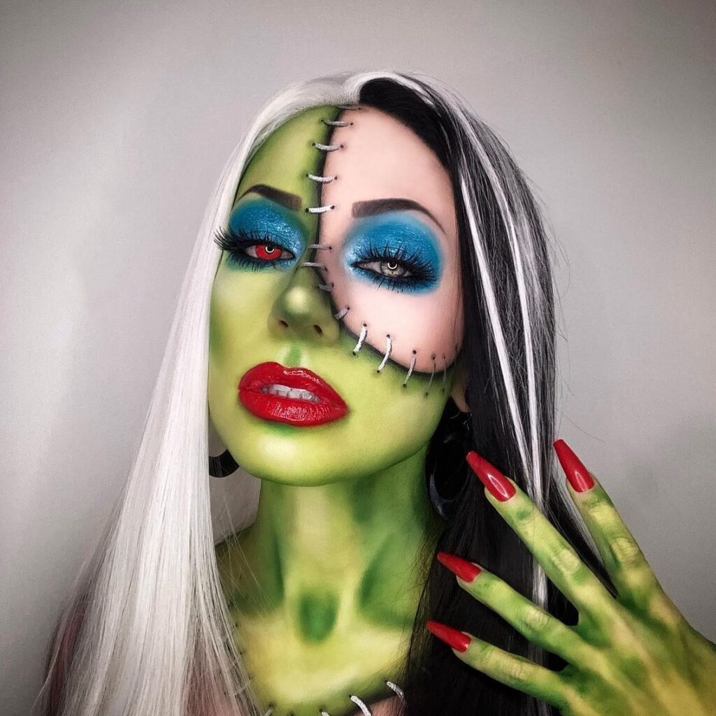 A person with green Frankenstein-inspired makeup, bold blue eyeshadow, red lips, and black stitches across the face, featuring two-toned black and white hair.