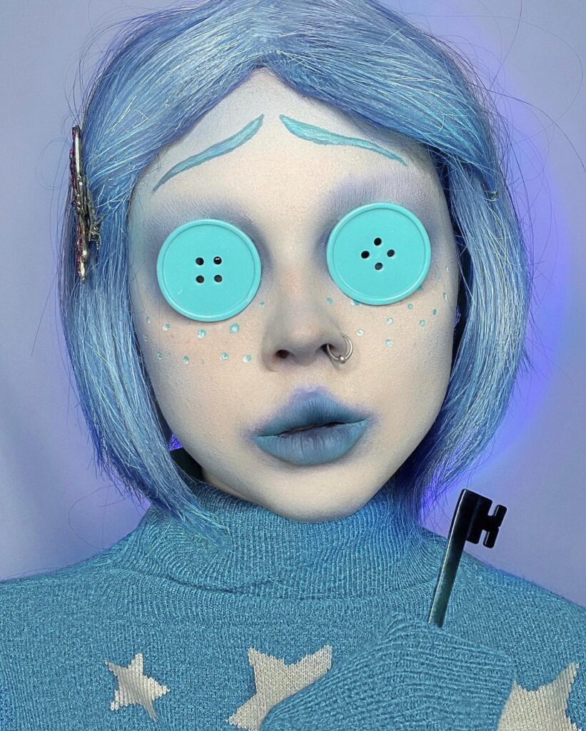 A person with pale blue makeup, button eyes, blue lips, and blue hair, holding a black key for a creepy doll-like appearance.