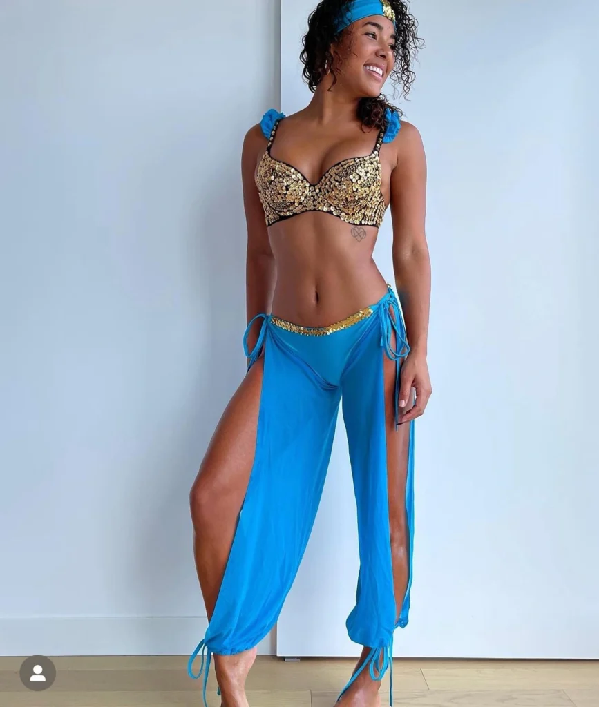 A woman wearing a genie-themed costume with a gold-sequined bra, blue harem pants with high slits, and a matching blue headband, smiling and posing in a bright room.