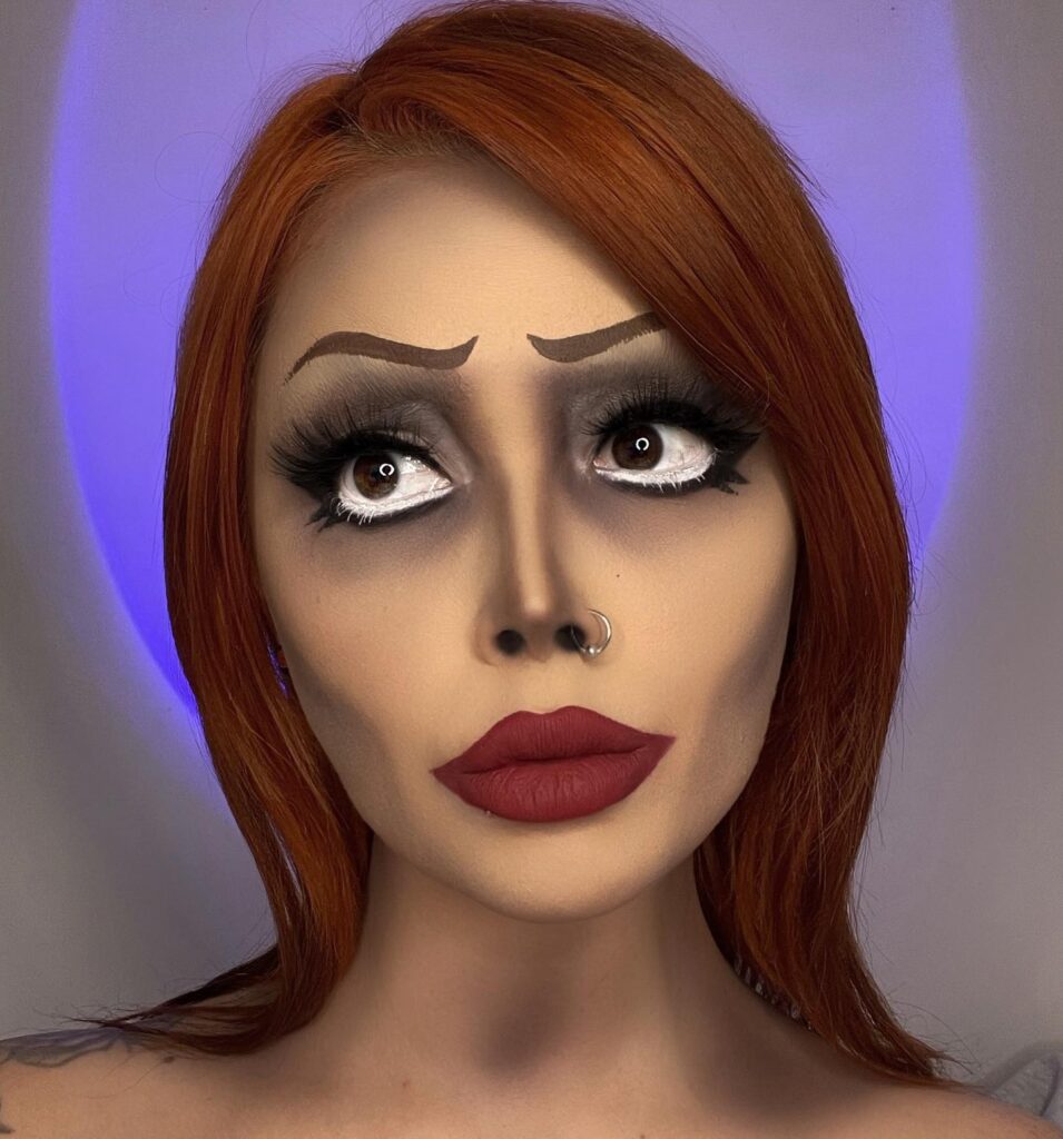 A person with animated doll-inspired makeup, featuring large, exaggerated eyes with bold shadows, red lips, and bright red hair.