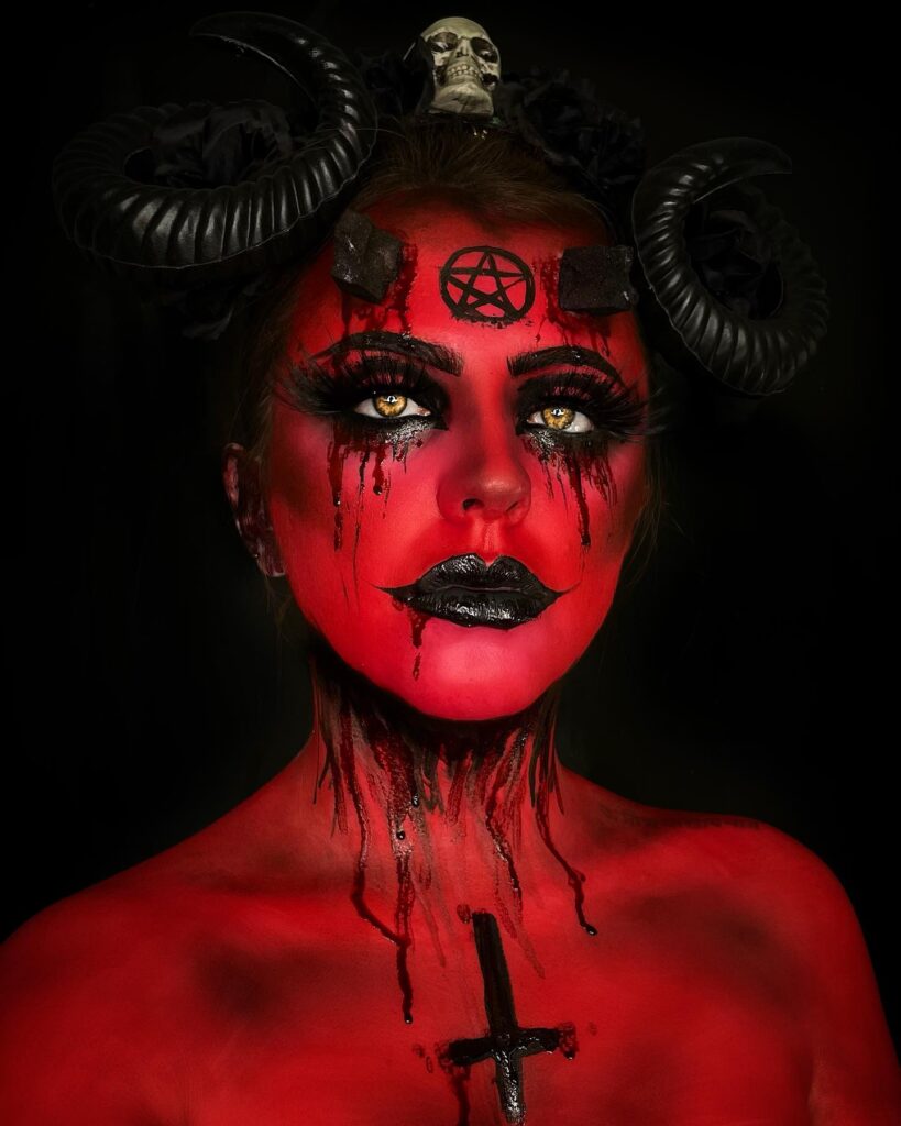 A person with red body paint, black horns, and a pentagram on the forehead, wearing bold black eye makeup and lips, creating a demonic devil look.