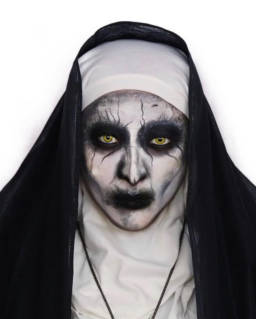 Person wearing demon nun-inspired Halloween makeup with cracked white skin, black eyeshadow, yellow contacts, and black lips, styled with a black and white nun's habit for a haunting look.