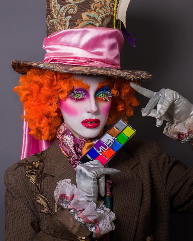 A person dressed as the Mad Hatter, featuring orange curls, a large patterned hat, rainbow makeup, and bold pink blush with glittering red lips.