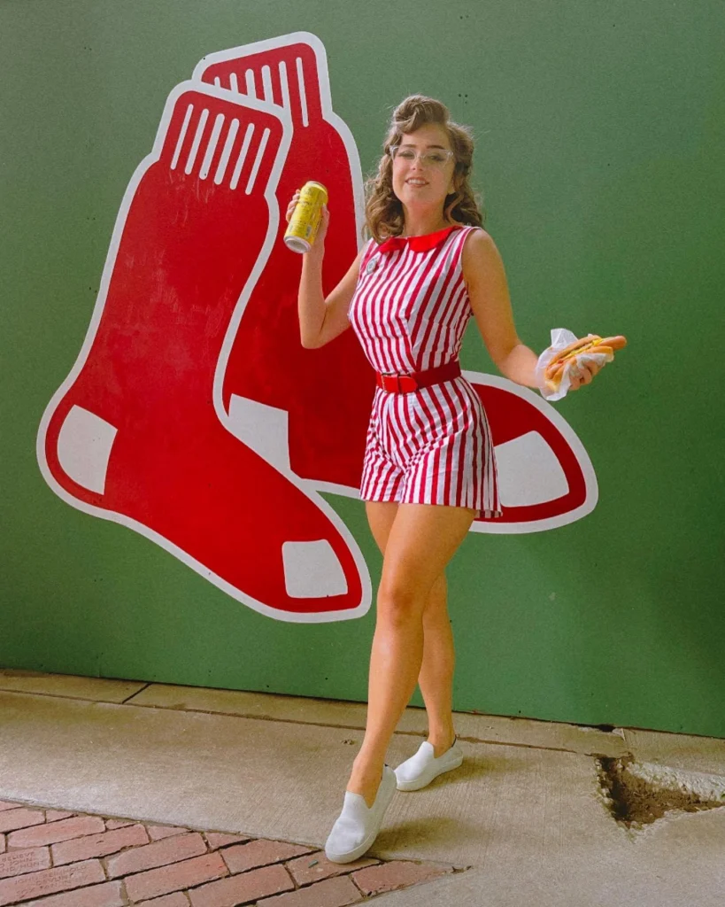 Woman dressed in a red and white striped romper with a red belt, white slip-on sneakers, and retro glasses, holding a hot dog and drink in front of a ballpark mural.