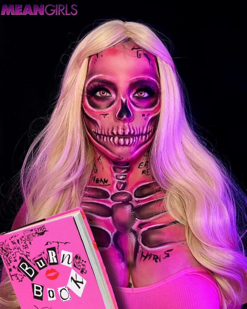 Person wearing a Mean Girls-inspired Halloween makeup look with a pink skeleton design, blonde wig, and holding a 'Burn Book' prop for a glamorous yet spooky effect.