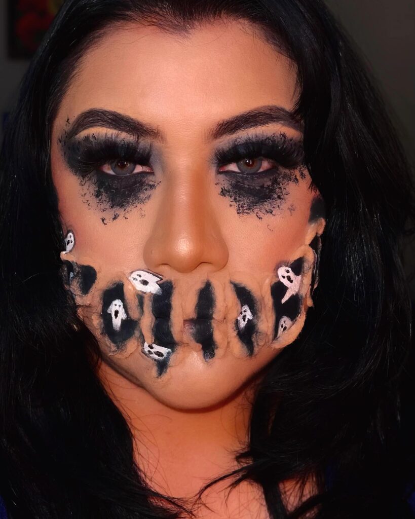 Person wearing ghostly Halloween makeup with a stitched mouth design featuring small ghost figures, bold black eyeshadow, and dramatic lashes for a haunting effect.