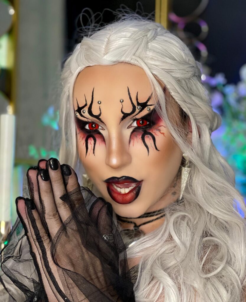 A person with silver hair and intense red eye makeup featuring flame-like black designs, paired with bold red lips, wearing a black lace glove, exuding a fierce sorceress vibe.