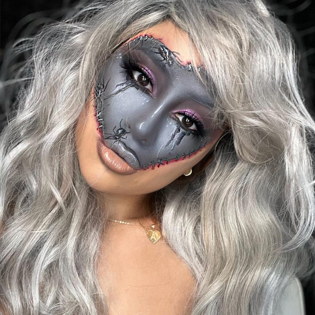 Person wearing stitched doll Halloween makeup with a grey face, sewn seams, smoky eyes, nude lips, and silver wavy hair for a creepy yet stylish look.