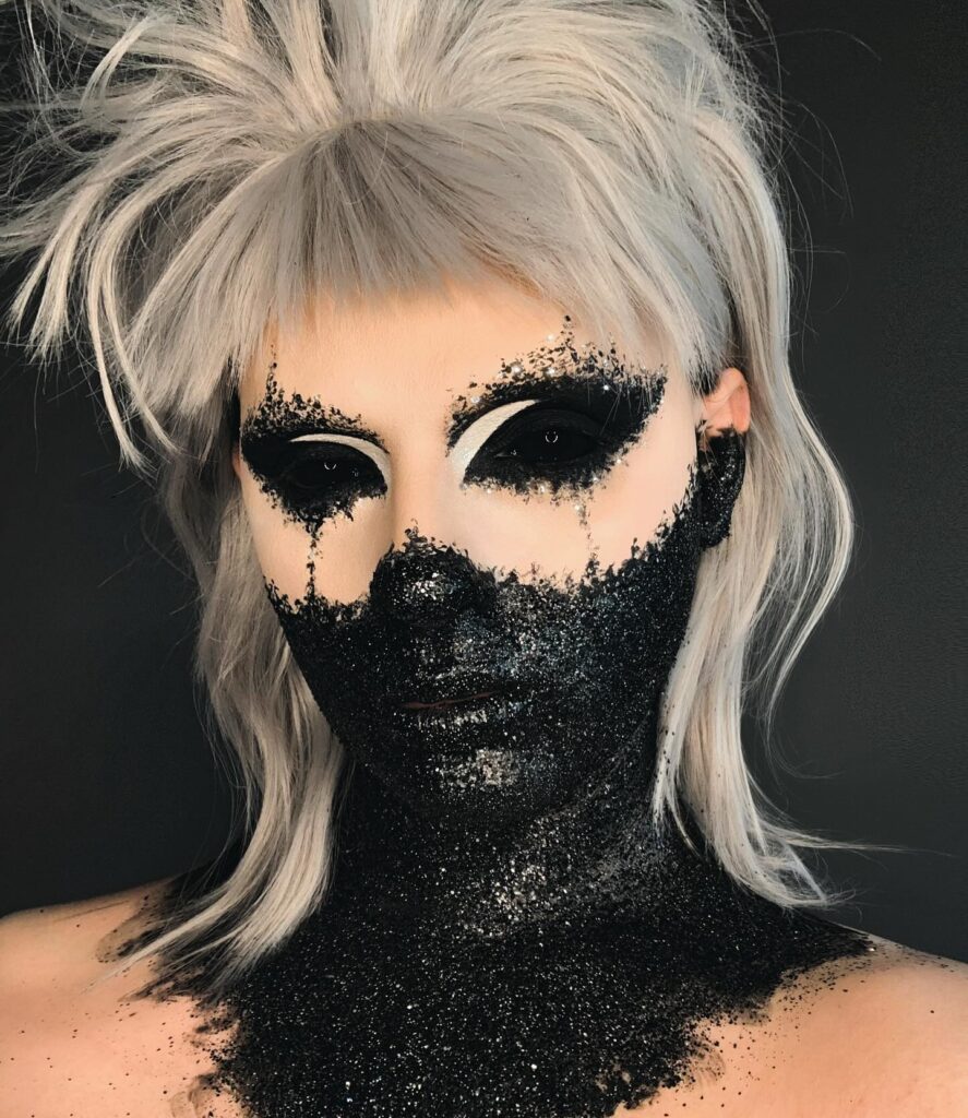 A person with silver hair and black glitter covering the lower face and neck, with dramatic black and white eye makeup creating a stark, otherworldly look.