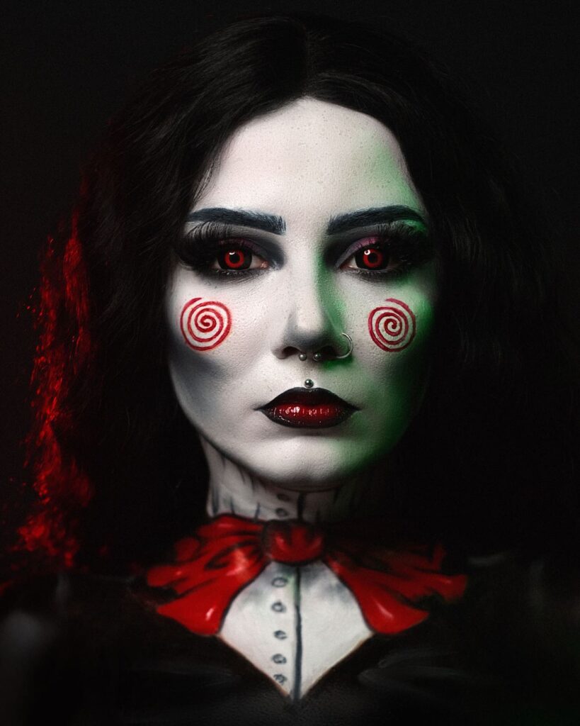 Person wearing spiral doll-inspired Halloween makeup with red spiral cheeks, red eyes, and painted red bow tie, styled to mimic a haunting doll aesthetic.