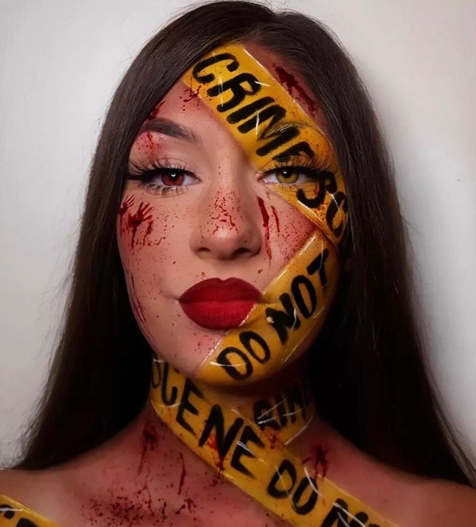 A creative Halloween makeup design featuring painted yellow "crime scene" tape across the face, splattered blood, and red handprints, paired with a bold red lip and glam eye makeup for a striking mix of horror and beauty.