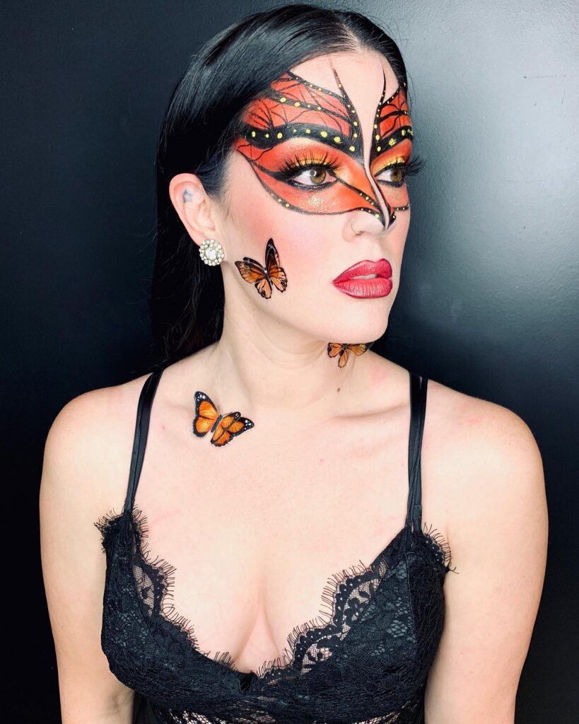 Person with monarch butterfly-inspired Halloween makeup featuring orange, black, and yellow eye makeup that extends into a butterfly wing design. Additional butterfly accents are placed on the cheek and collarbone.