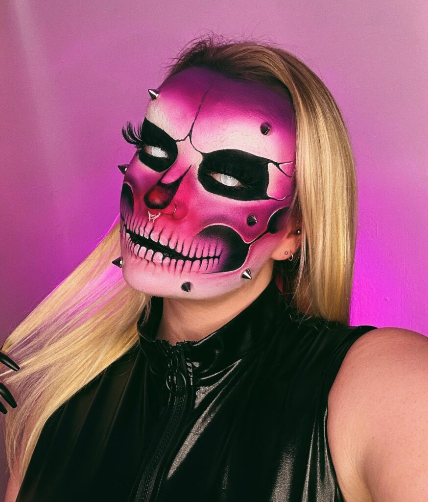 A person with neon pink and purple skull makeup, featuring black contouring and spikes embedded in the skin, paired with long eyelashes and black nails.