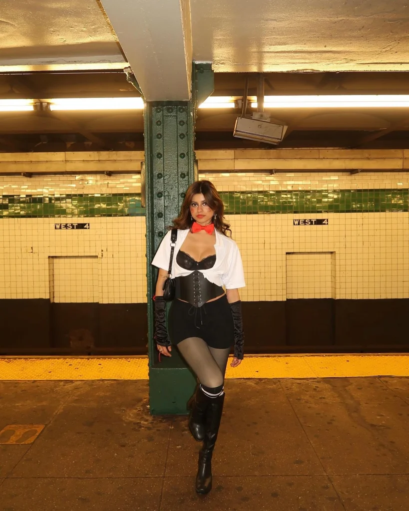 Woman in a black corset and white cropped shirt, styled with a red bowtie, high-waisted shorts, and knee-high boots, standing in a subway station.