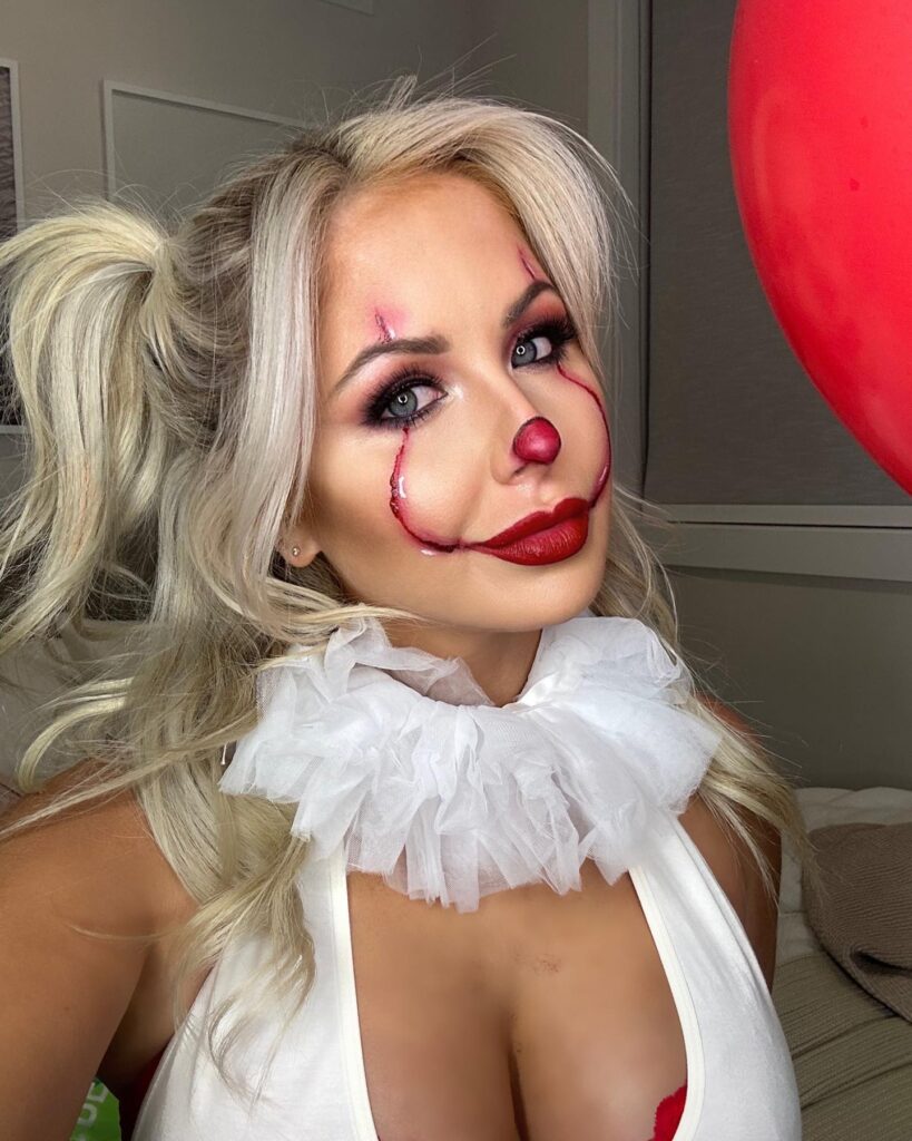 A woman with white-blonde hair styled in ponytails, wearing red clown makeup with a white ruffled collar, inspired by Pennywise.