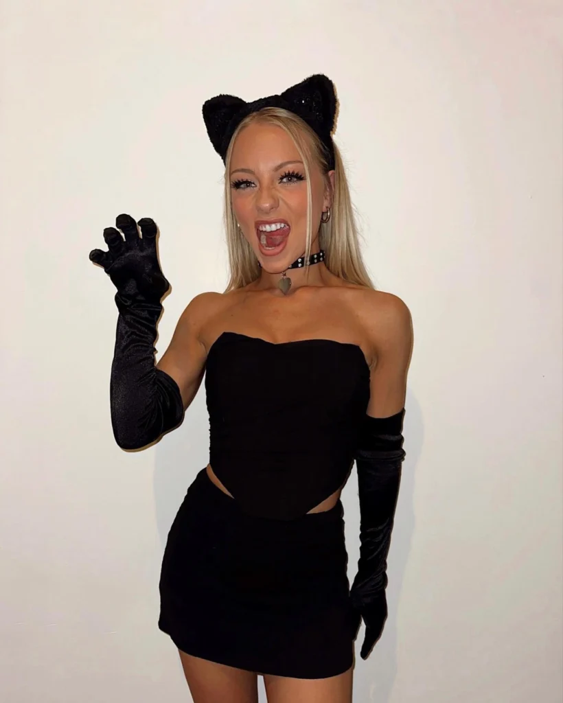 Woman dressed in a black strapless top and mini skirt, accessorized with cat ears, long gloves, and a studded choker, posing with a playful cat-like expression."