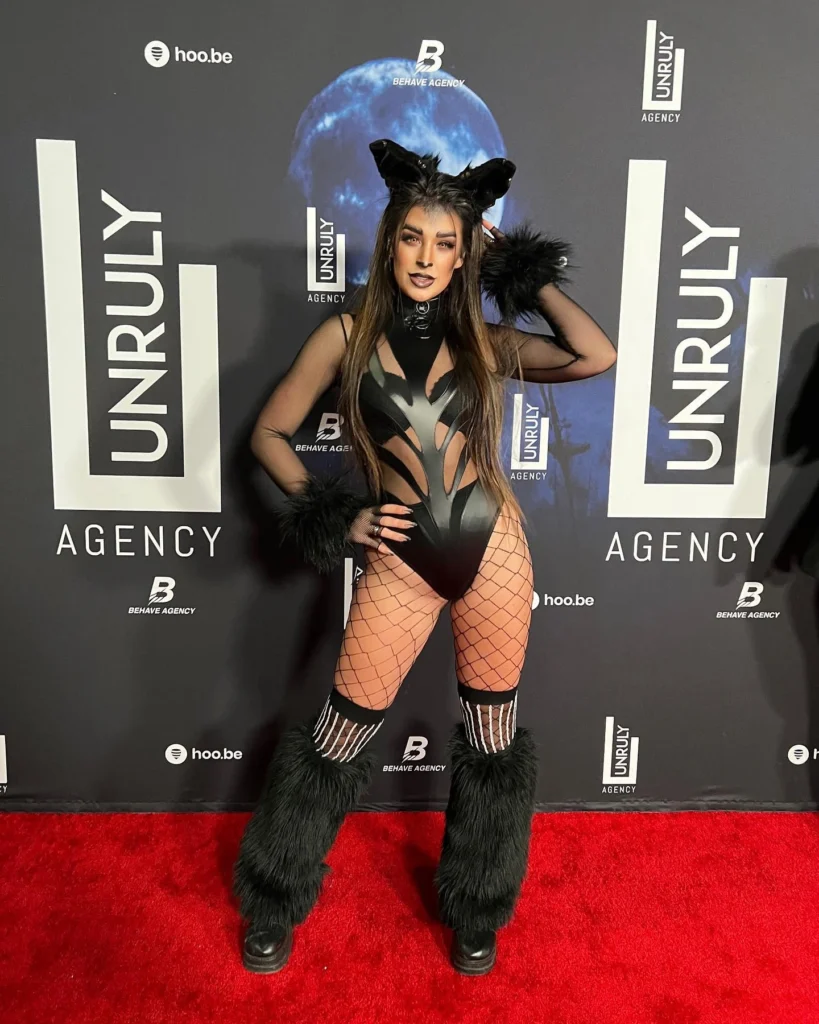 Woman in a fierce feline costume with a black cutout bodysuit, fishnet stockings, furry leg warmers, and cat ears, posing on the red carpet.