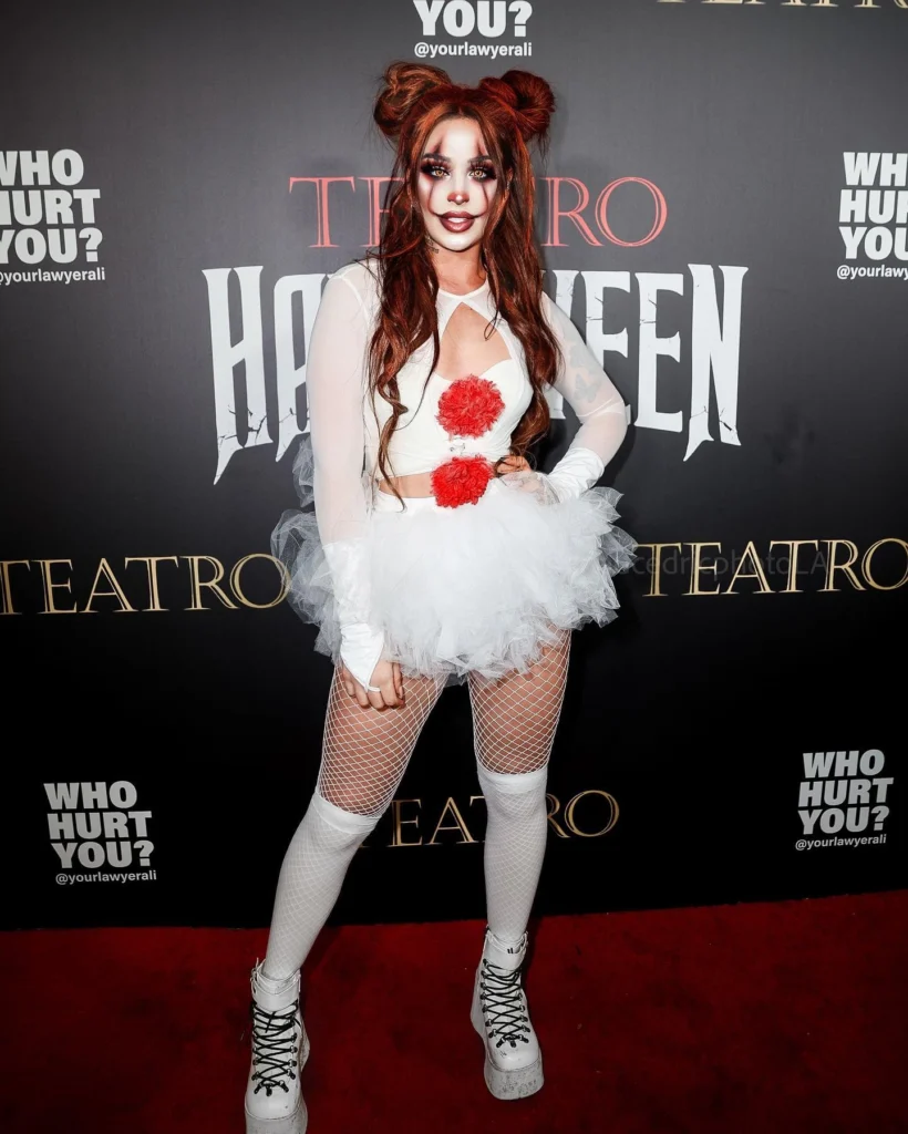 Woman in a whimsical horror clown costume with a white tutu dress, red pompoms, fishnet stockings, and white platform boots, standing on the red carpet.