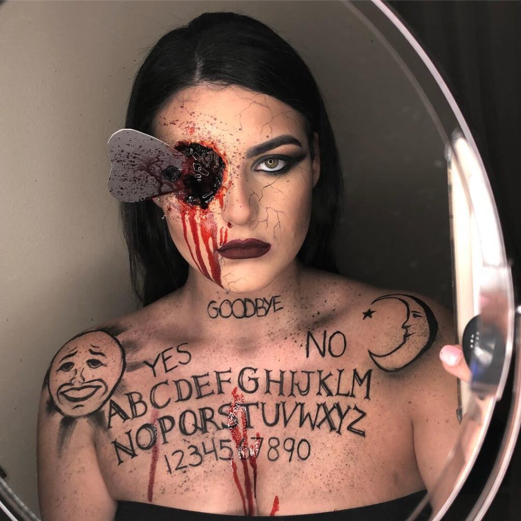 Person wearing Ouija board-themed Halloween makeup with a bloody planchette embedded in the eye and Ouija board letters and symbols painted across the chest for a terrifying, supernatural look.