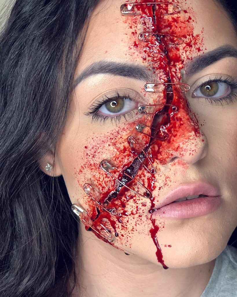 Close-up of a woman's face featuring special effects makeup with safety pins and blood creating the illusion of a stitched wound running across her face.